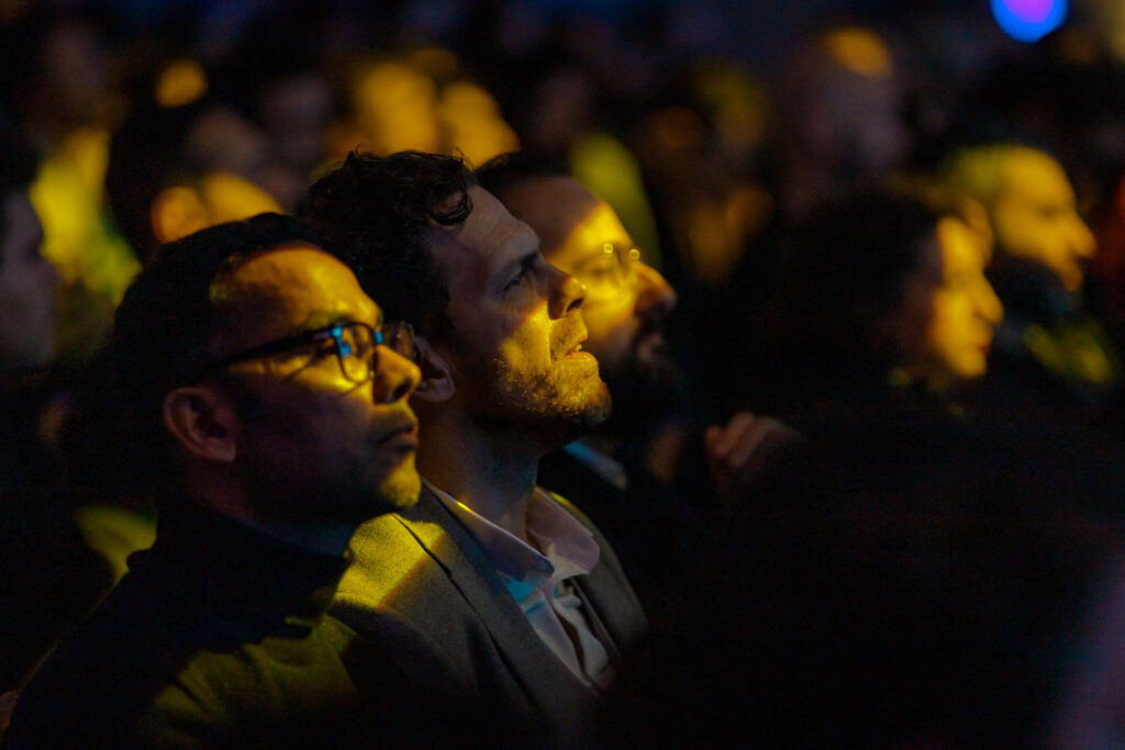 Audience members focused, illuminated by yellow stage light.
