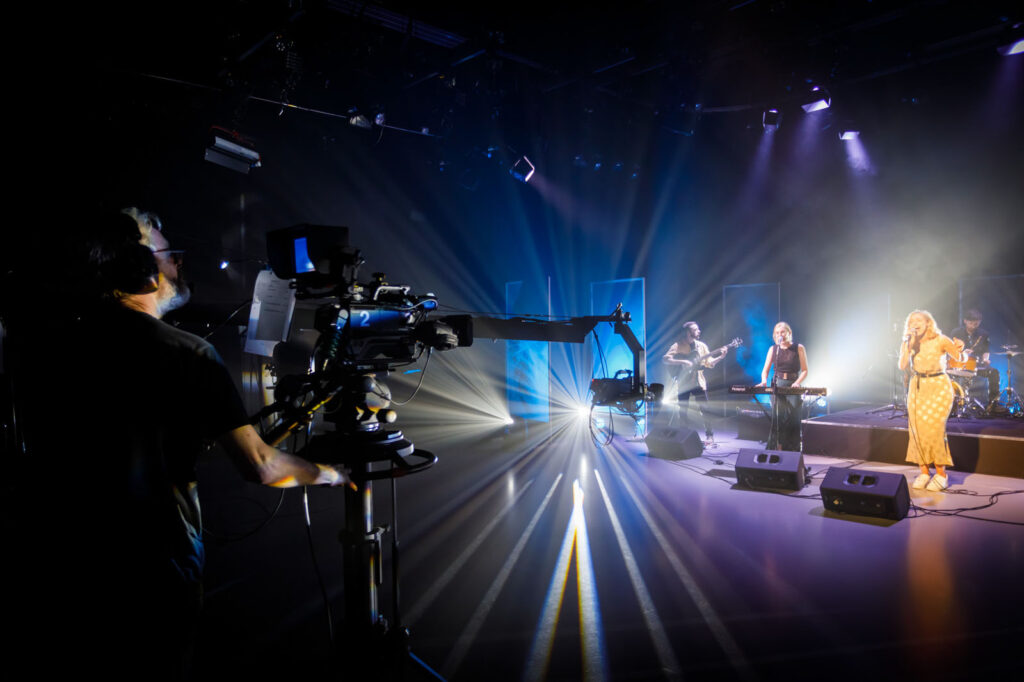 Band performs live on stage, filmed by a camera.