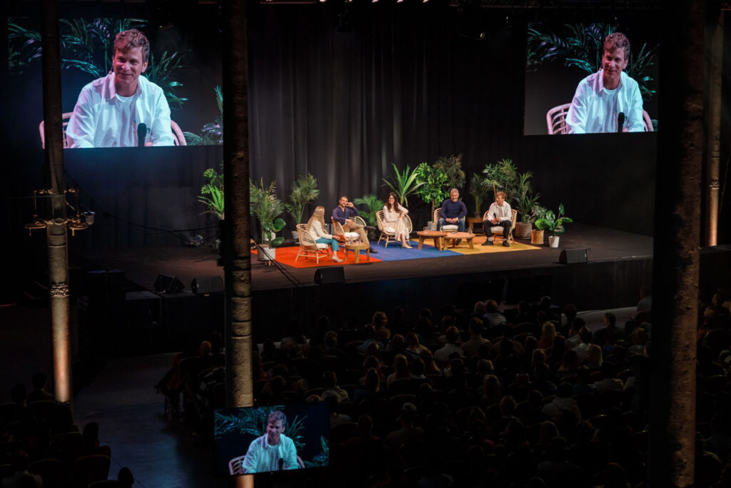 Conference panel discussion with speakers, audience, and large screens.