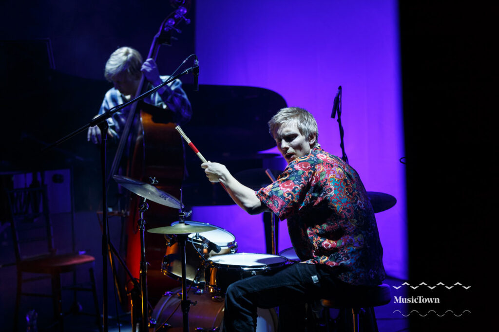 Drummer and bassist performing live on a purple-lit stage.