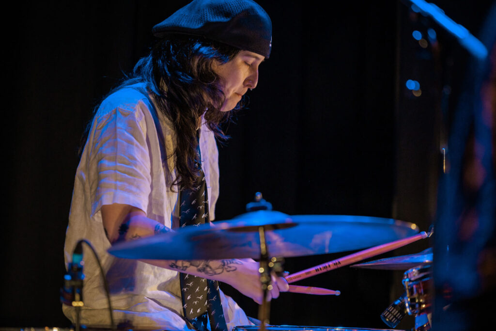 Drummer in hat and dinosaur tie playing under blue light.