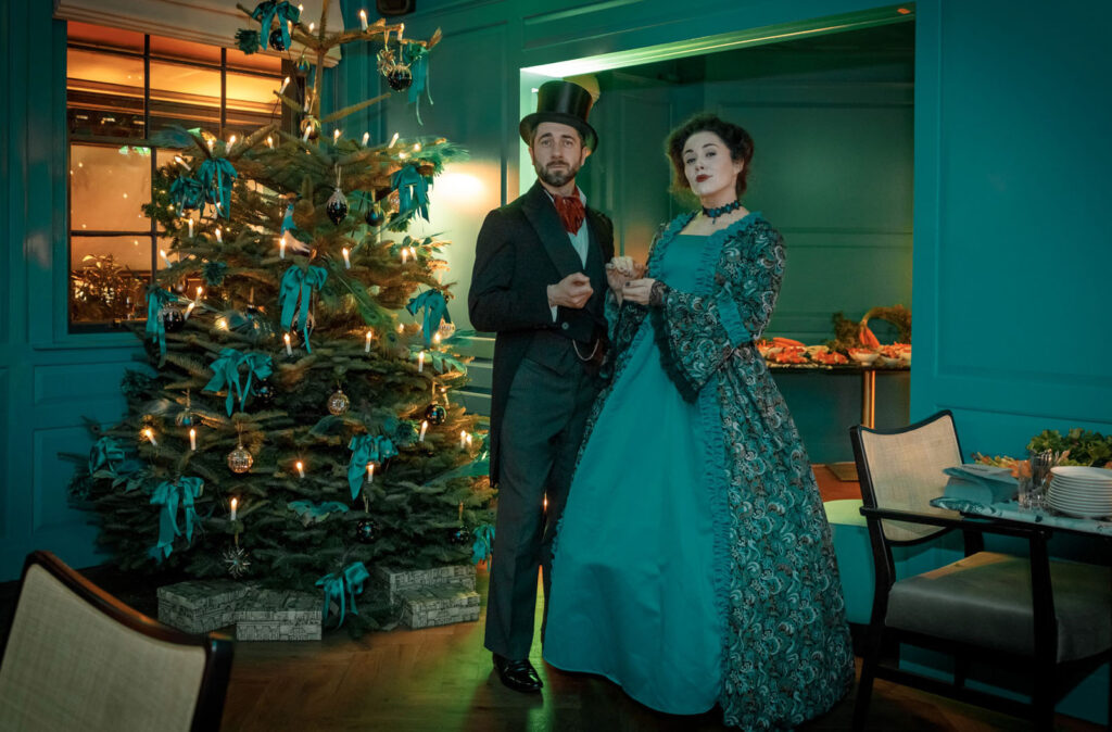 Elegant couple in period attire by a decorated Christmas tree.