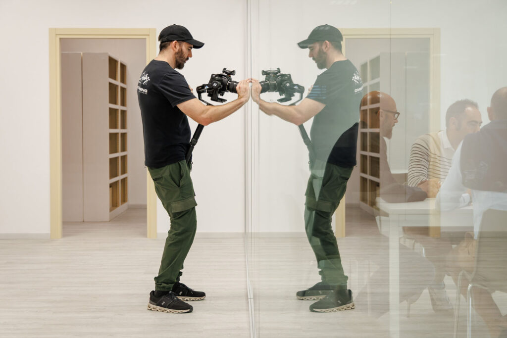 Filmmaker with camera rig reflected in glass, people visible beyond.