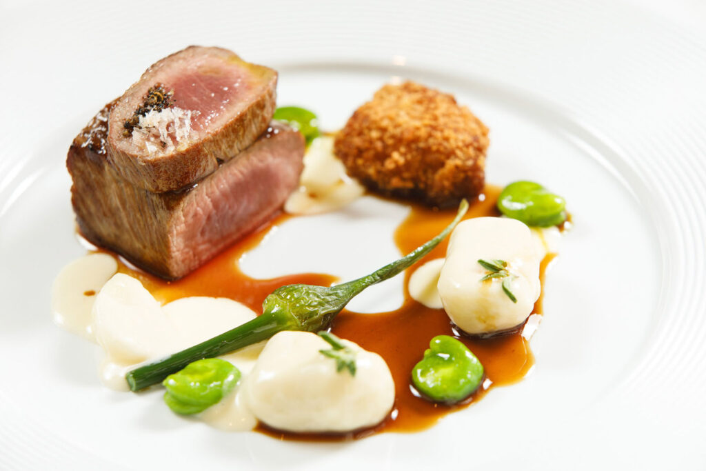 Gourmet dish with seared meat, croquette, vegetables, and sauce.