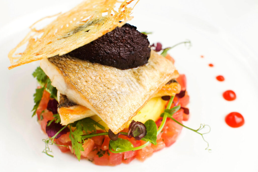 Gourmet fish dish with dark topping, crispy garnish, and tomato base.