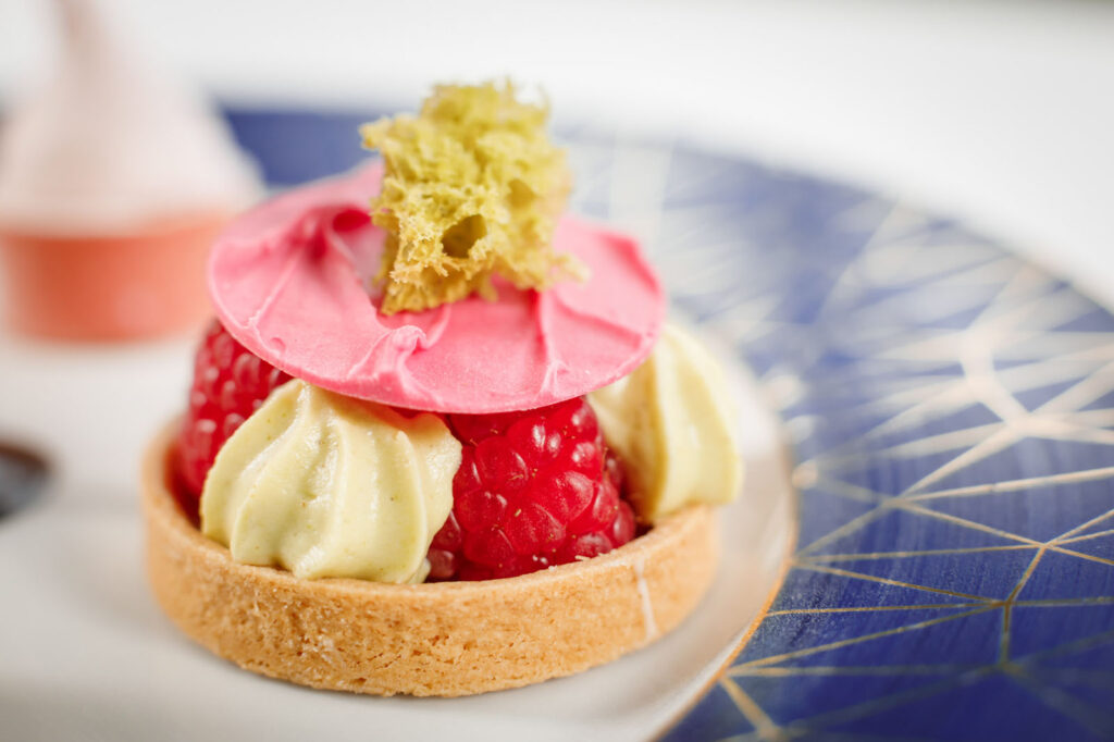 Gourmet raspberry tart with pistachio cream and pink chocolate decoration.