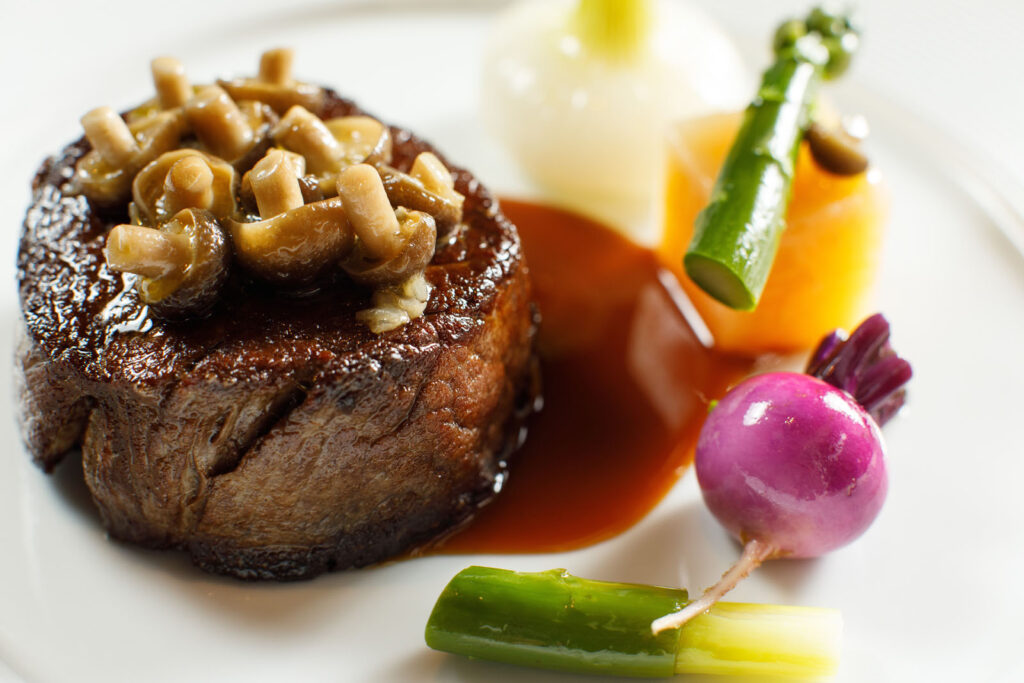 Gourmet steak with mushrooms and colorful vegetables on a white plate.