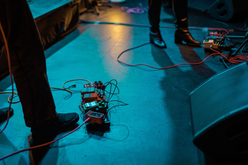 Guitar pedals and musician's feet on a blue-lit stage.