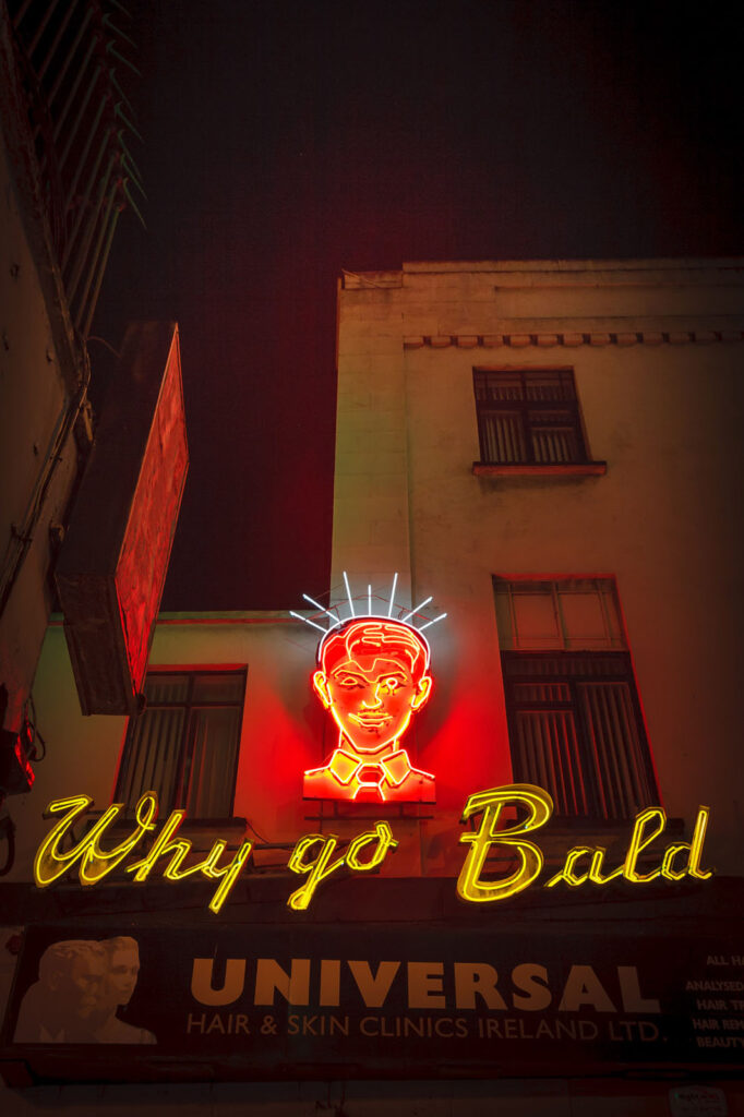 Hair clinic's 'Why go Bald' neon sign glows at night.