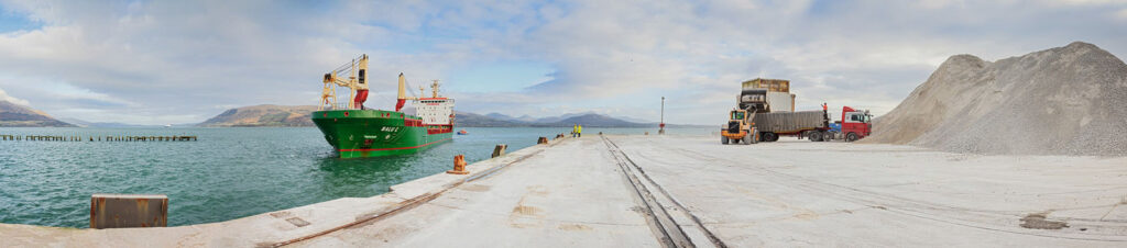 Industrial port with ship, gravel, and mountains.