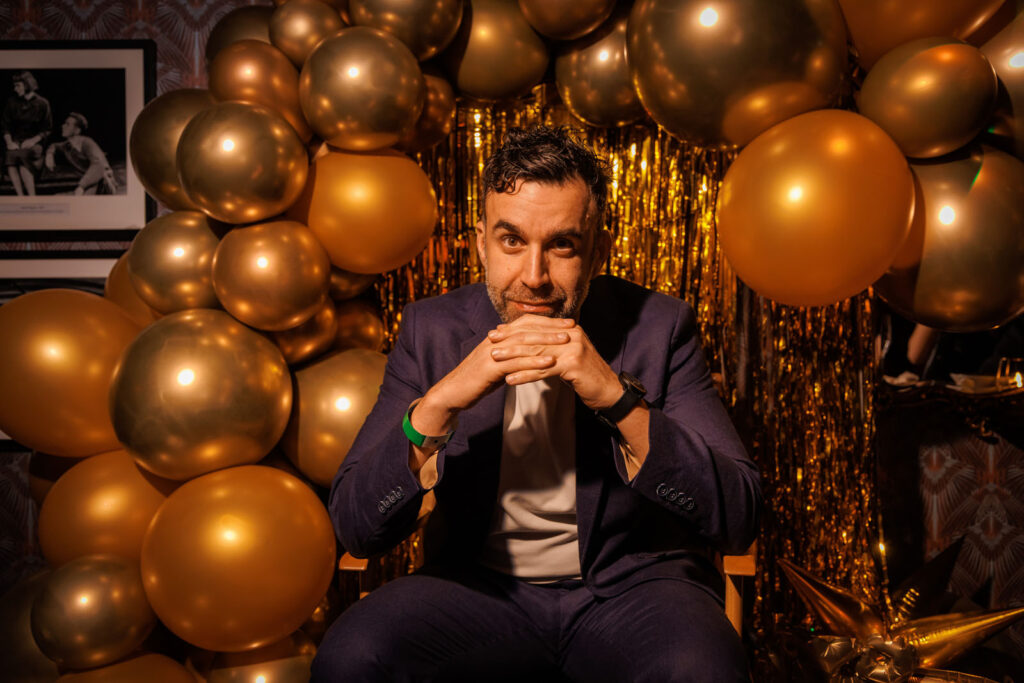 Man in suit poses with golden balloons and tinsel.
