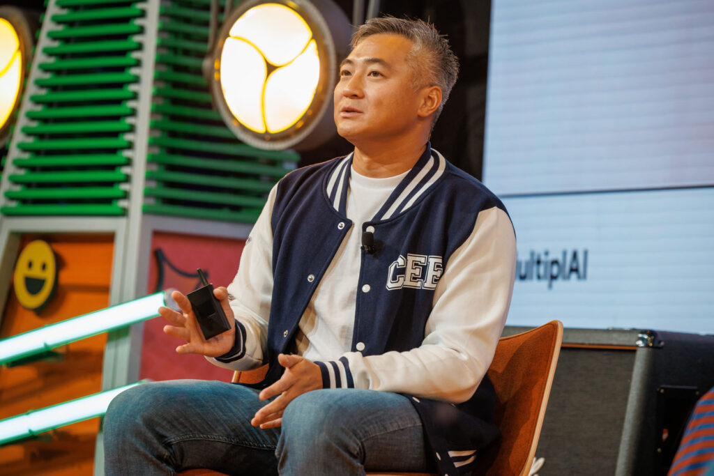 Man in varsity jacket speaks on stage, holding device.