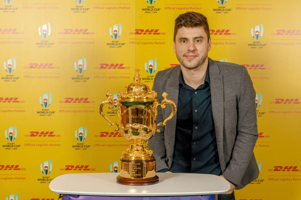Man poses with Rugby World Cup trophy, DHL branding.