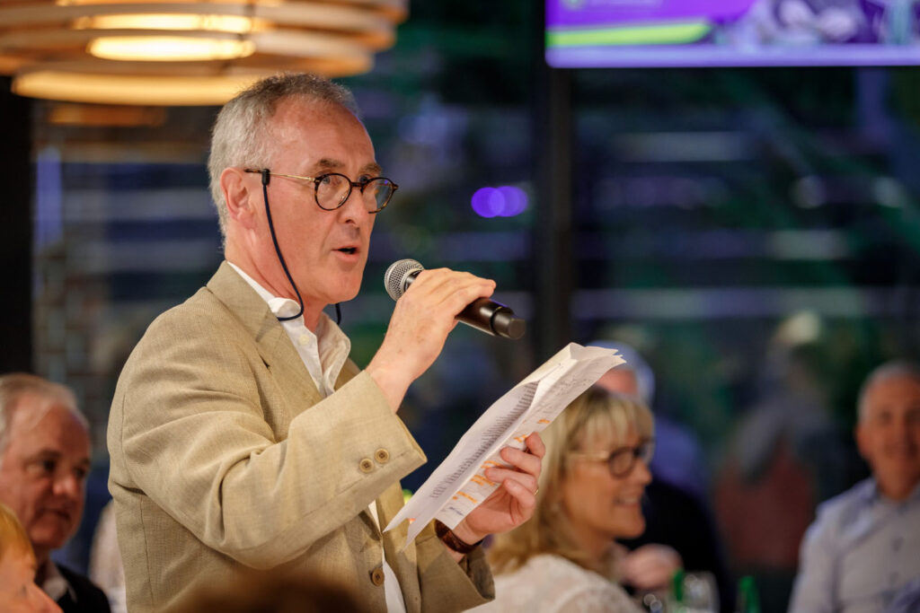 Man speaking, holding microphone and notes.