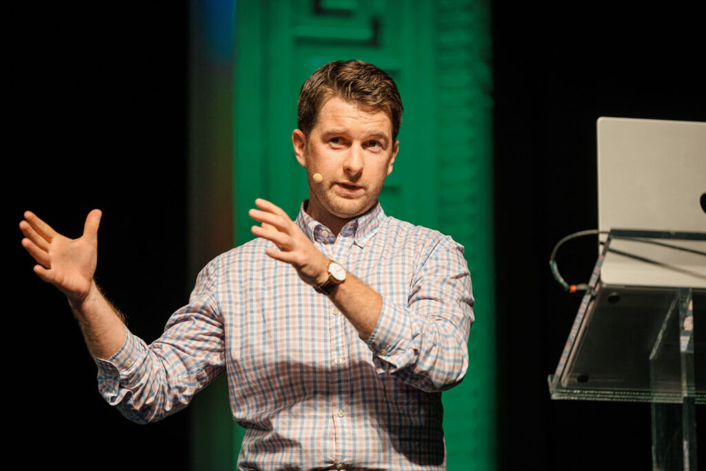 Man speaking on stage with laptop and green background.