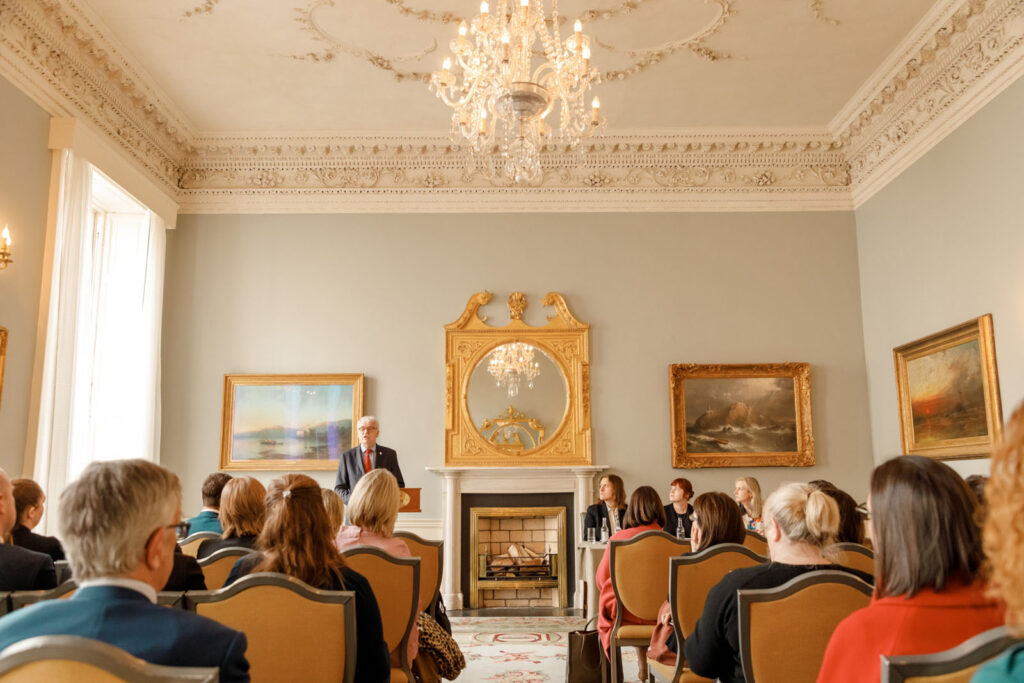 Man speaks to audience in elegant room.