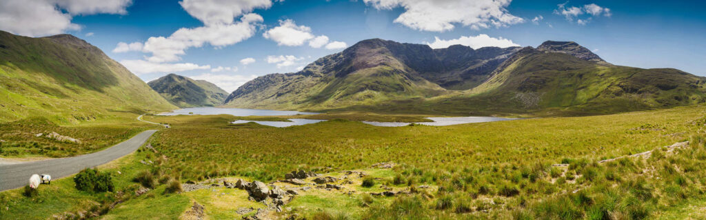 Panoramic view of green mountains, lake, road, and sheep.
