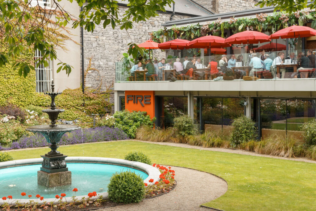 Restaurant balcony with diners, red umbrellas, and garden fountain.