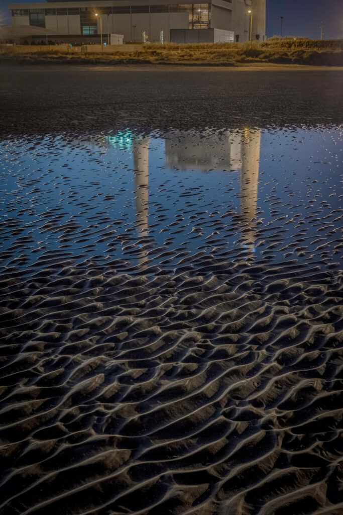 Rippled sand reflects industrial building at night.