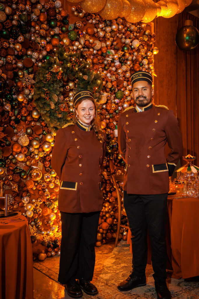 Smiling bellhops in uniform against a festive Christmas ornament wall.