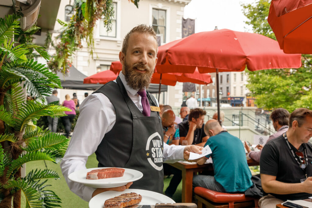 Smiling waiter serves steak at an outdoor dining event.