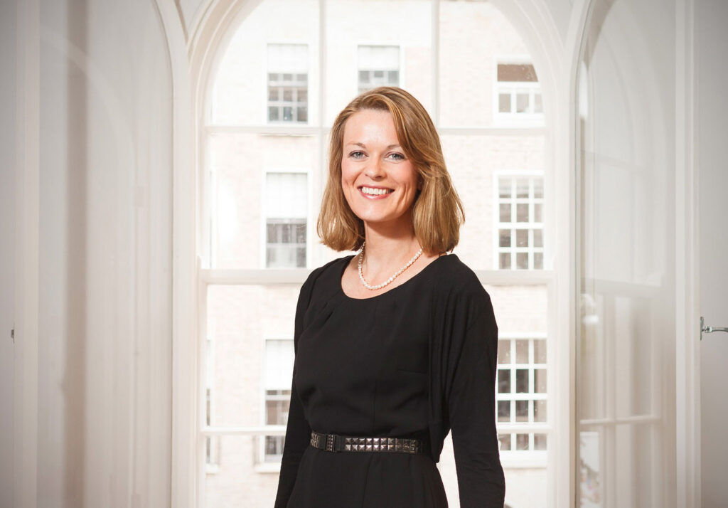 Smiling woman in black dress and pearls by a window.