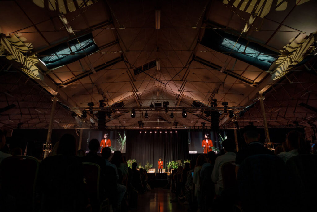 Speaker addresses audience at a large, dimly lit conference.