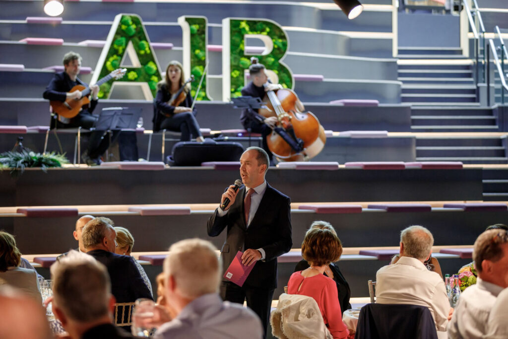 Speaker addresses audience, musicians perform under "AIB" sign.