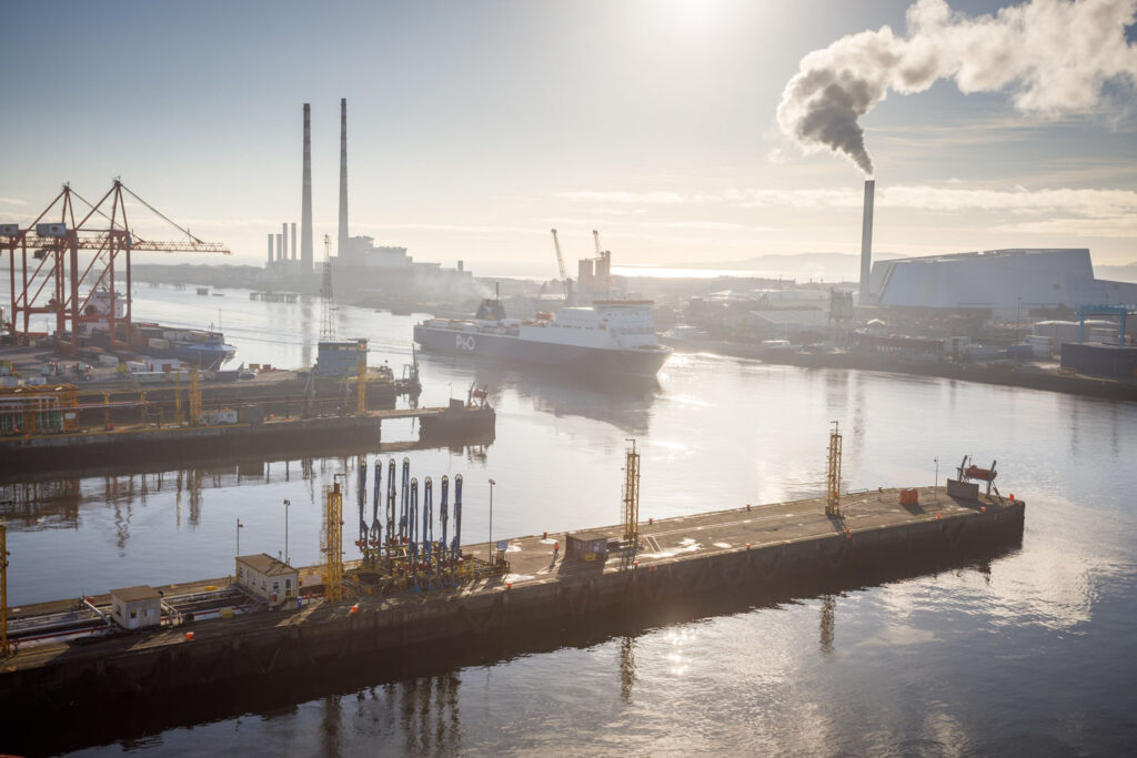 Sunlit industrial port with ferry, cranes, and smoking chimneys.