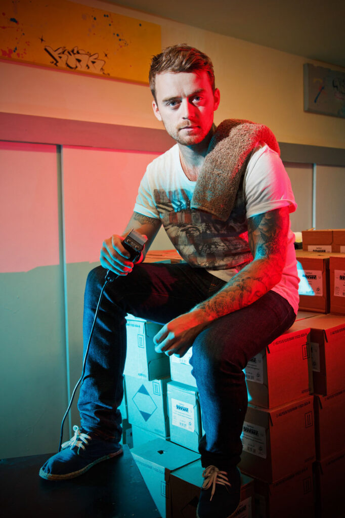 Tattooed man with clippers sits on boxes in colorful lighting.