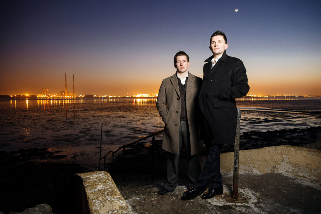 Two men in coats by waterfront with city lights at dusk.