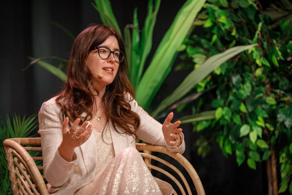 Woman in glasses speaking, gesturing, with lush green background.