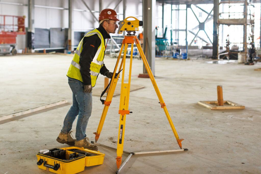 Worker surveying construction site with leveling instrument.